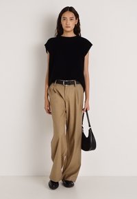 Czarny top z krótkim rękawem w zestawieniu z beżowymi spodniami z wysokim stanem, czarnym paskiem i czarną torebką. Modelka stoi na neutralnym tle.