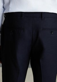 Pantalon marine en tissu lisse, présentant deux poches arrière avec fermeture à bouton et une coupe ajustée. Motif rayé visible sur la chemise.