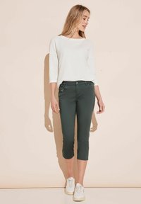 Pantalones recortados verdes de corte ajustado, con cierre de botón y detalle en los bolsillos laterales, combinados con una blusa blanca de manga larga y corte holgado.