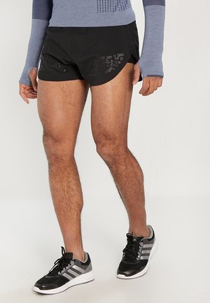 Homme portant un short de sport noir avec des découpes à motifs, un haut à manches longues gris avec des trous pour les pouces, et des chaussures de course Adidas noires et grises.