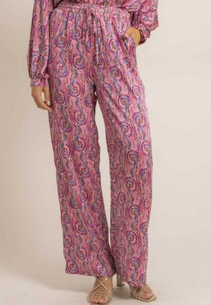 Femme portant un pantalon large à motifs roses avec une taille à cordon et des sandales à talons hauts métalliques à brides, une main dans la poche.