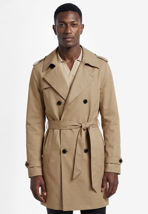 Man draagt een beige trenchcoat met dubbele rij knopen, ceintuur gestrikt in de taille en zwarte knopen, staand voor een witte achtergrond.