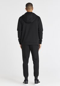 Sudadera negra con una gran capucha y mangas largas, combinada con pantalones de chándal a juego. Tela suave, costuras mínimas, diseño ajustado y puños acanalados.
