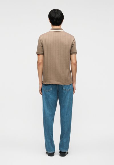 Άντρας όρθιος, γυρισμένος με την πλάτη, φοράει καφέ πλεκτό πόλο με κοντά μανίκια και μπλε τζιν με ίσια πόδια, μαζί με μαύρα παπούτσια, σε λευκό φόντο.