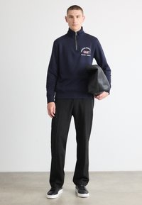 Navy zip-up sweatshirt with logo, paired with black trousers and black sneakers. The model holds a black textured bag. Smooth fabric texture.