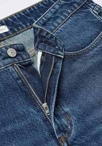 Jean en denim avec une braguette zippée, de couleur bleu foncé, coutures orange et fermeture par bouton en métal. La texture semble lisse avec un léger délavage.