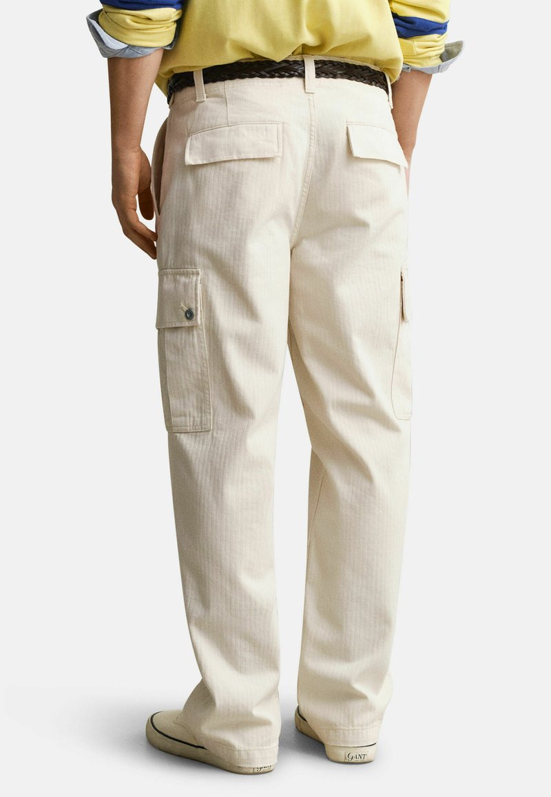 Personne portant un pantalon cargo couleur crème avec des poches à rabat, une ceinture tressée marron, une chemise jaune et des baskets claires, debout de dos face au spectateur.