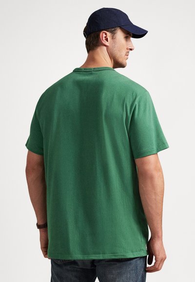 Vyras dėvi žalią trumpomis rankovėmis marškinius, tamsiai mėlyną kepurę, mėlynas džinsus ir riešo laikrodį, žiūri į priešais esančią paprastą baltą foną.