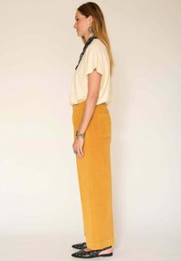 Haut beige à manches courtes, pantalon large en velours côtelé jaune avec une poche arrière, ballerines noires à bout pointu avec des accents ronds, et une écharpe noire à motifs.