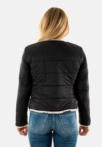 Chaqueta acolchada negra con costuras horizontales, que presenta un borde suave blanco en el dobladillo y los puños. Combinada con jeans azules. Vista desde atrás.