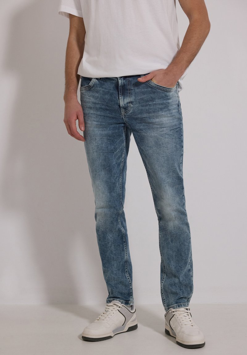 Homme portant un jean bleu délavé, des baskets blanches et un t-shirt blanc, une main dans la poche, debout devant un fond uni.