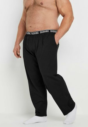 Shirtloze man die zwarte loungebroeken met elastische tailleband en witte sokken draagt, staand tegen een effen lichte achtergrond.