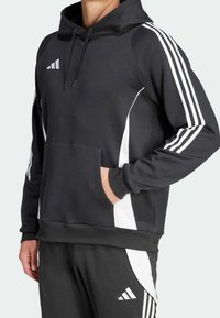 Uomo che indossa un hoodie Adidas nero e pantaloni abbinati con strisce bianche e logo, con una mano nella tasca.