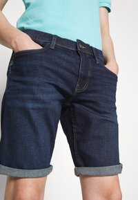 Shorts en denim bleu foncé avec une finition légèrement délavée, présentant des ourlets retroussés et un design classique à cinq poches.