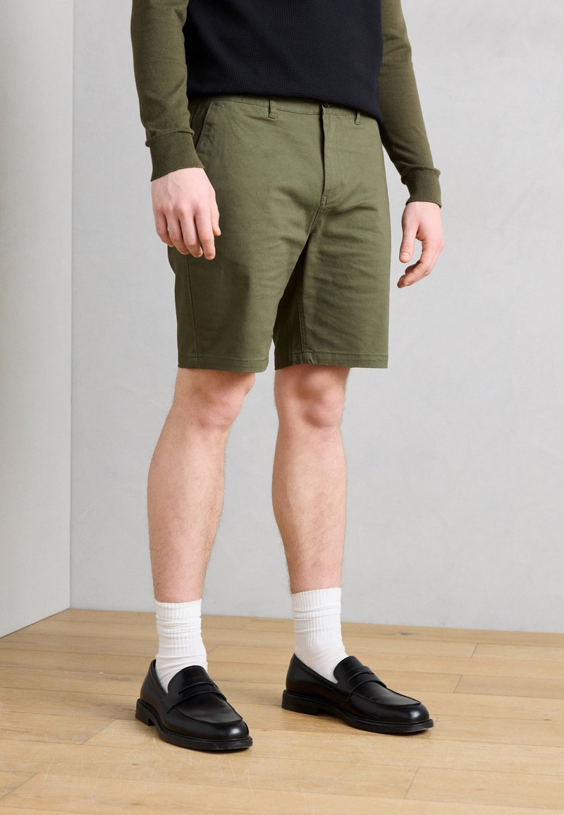 Άνδρας που φοράει χακί πράσινα σορτς, μαύρα μοκασίνια, λευκές κάλτσες και χακί πράσινο πουκάμισο με μακριά μανίκια και μαύρο γιλέκο στέκεται σε ξύλινο πάτωμα.