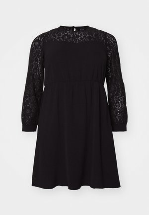 Vestido preto com um sobrepano de renda nas mangas longas, gola redonda e uma cintura elástica, apresentando uma silhueta solta e fluida.