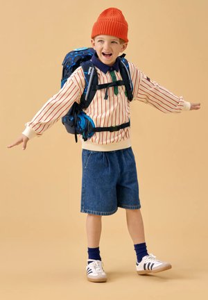 Young boy wearing orange beanie, striped sweater, denim shorts, and sneakers with a large blue backpack, arms outstretched, smiling.