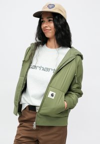 Olive zelena jakna s kapuco, zadrga spredaj, rebrasti manšeti, stranske žepe, tekstura tkanine. Nosi se preko sive majice s kapuco z napisom "Carhartt".