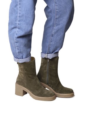 Stivaletti alla caviglia in suede verde oliva, con un tacco rotondo spesso, zip laterale e suola testurizzata in contrasto. Indossati con jeans di denim arrotolati.