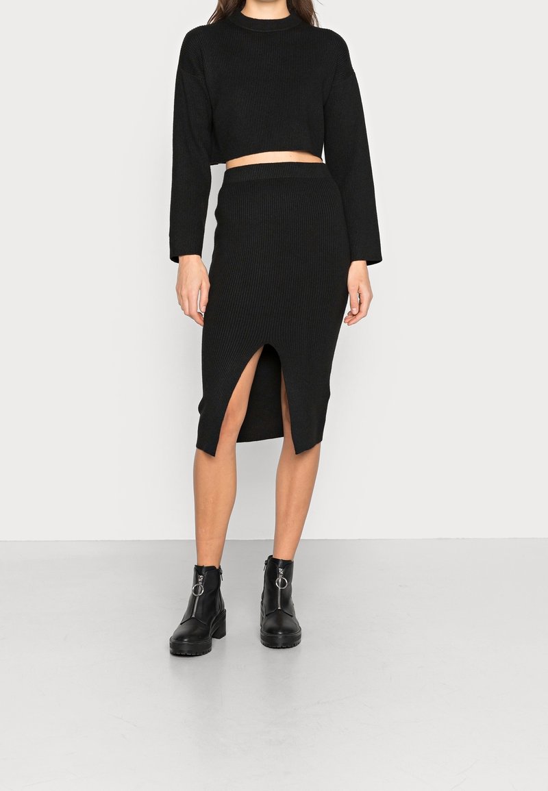 Femme portant un crop top noir côtelé manches longues et une jupe midi taille haute assortie avec une fente devant, associée à des bottines noires.