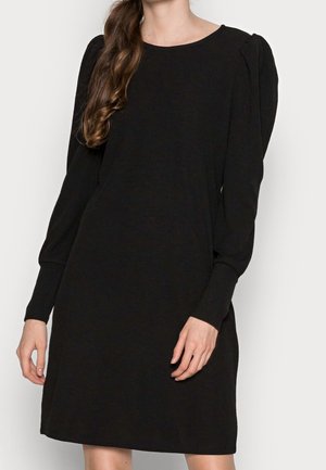 Kobieta nosząca czarną sukienkę z długim rękawem z bufkami na ramionach i dopasowanymi mankietami, stojąca na tle jednolitego białego tła.