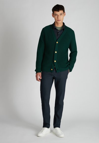 Zielony sweter z dzianiny o teksturowej tkaninie, z dekoltem w kształcie litery V i trzema złotymi guzikami. Noszony na ciemnej koszuli, zestawiony z granatowymi spodniami.
