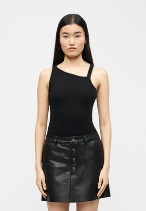 Γυναίκα με μακριά σκούρα μαλλιά που φοράει μαύρο τοπ με έναν ώμο και μαύρη κοντή δερμάτινη φούστα με κουμπιά μπροστά, στέκεται μπροστά σε ένα απλό φωτεινό φόντο.