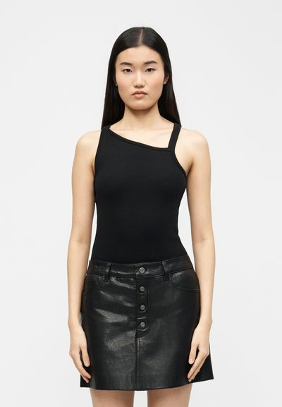 Femme aux longs cheveux noirs portant un haut noir à une épaule et une mini-jupe en cuir noire à boutons sur le devant, debout devant un fond clair uni.