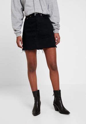 Personne portant un sweat à capuche gris, une mini-jupe en denim noire avec un ourlet effiloché et des bottines noires à talons, debout sur un fond blanc uni.