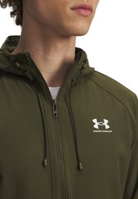 Olive spalvos gobtuvu atidaroma striukė, pagaminta iš lygaus audinio; turi priekinį užtrauktuką, raištelius ir baltą Under Armour logotipą ant krūtinės.