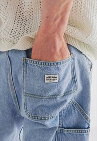 Pantaloni in denim azzurro chiaro con una grande tasca posteriore esterna che mostra un'etichetta bianca. Il tessuto presenta una texture e dettagli di cuciture visibili.