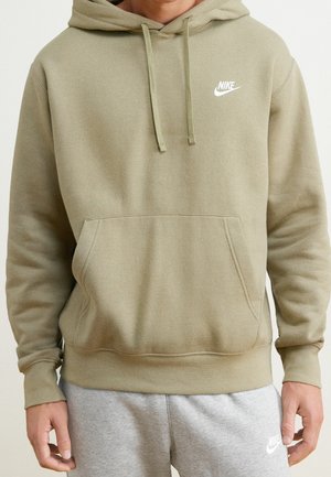 Mężczyzna noszący oliwkowo-zieloną bluzę Nike z kapturem i kieszenią na przodzie oraz szare spodnie dresowe Nike, stoi z rękami swobodnie opuszczonymi wzdłuż ciała.