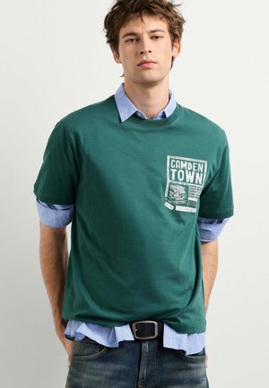 Giovane uomo con una maglietta verde sopra una camicia blu con bottoni, in piedi con le mani nelle tasche, rivolto in avanti su uno sfondo semplice.
