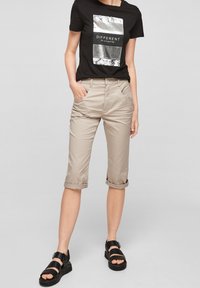Beige capri kalhoty s ohrnutými nohavicemi, v kombinaci s černým tričkem se stříbrným grafickým potiskem. Černé páskové sandály dokončují celkový vzhled.
