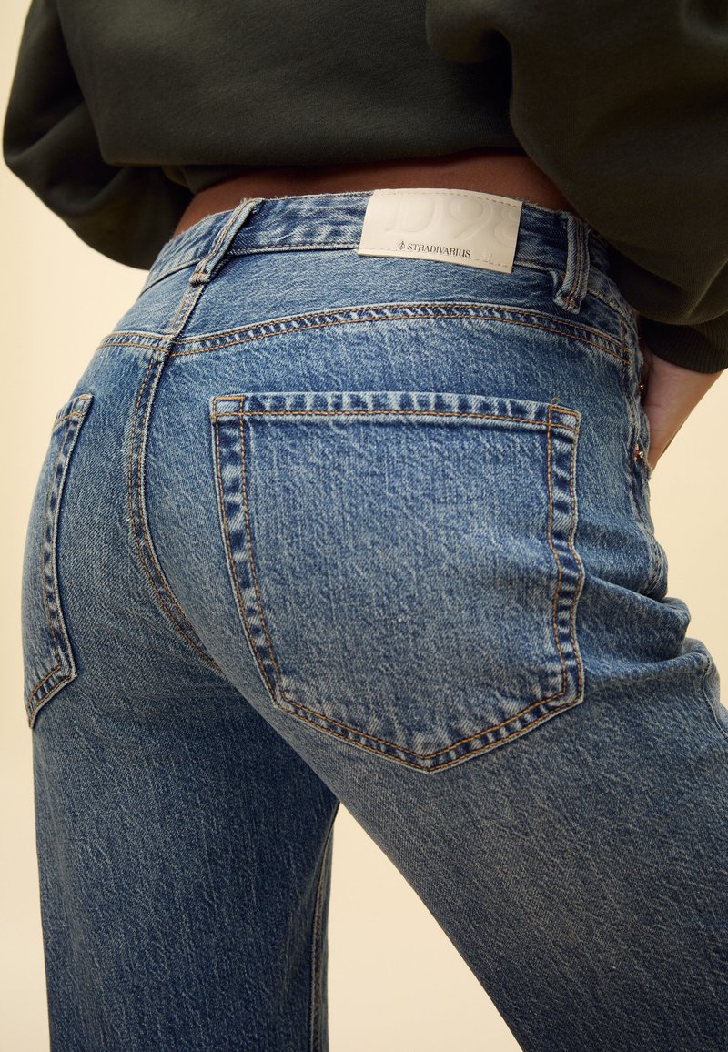 Primo piano di una persona che indossa jeans blu sbiaditi a gamba dritta con tasche posteriori e un'etichetta bianca visibile sulla cintura.