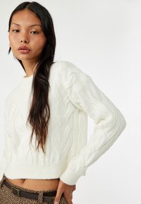 Maglione bianco a treccia con maniche lunghe e scollatura tonda, con polsini e orlo a coste. La texture è morbida, con un taglio leggermente corto.