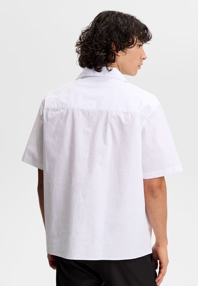 Mężczyzna o kręconych włosach, ubrany w luźną, białą koszulę z krótkim rękawem, zwrócony tyłem na jednolitym jasnym tle.