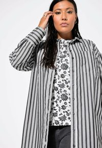 Camicia a righe bianche e nere con taschino anteriore, sovrapposta a un top a fantasia floreale, indossata con jeans scuri. Tessuto liscio.
