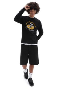 Sudadera negra con un estampado gráfico, combinada con pantalones cortos negros. El modelo lleva zapatillas negras y calcetines blancos. Cabello ligero y texturizado visible.