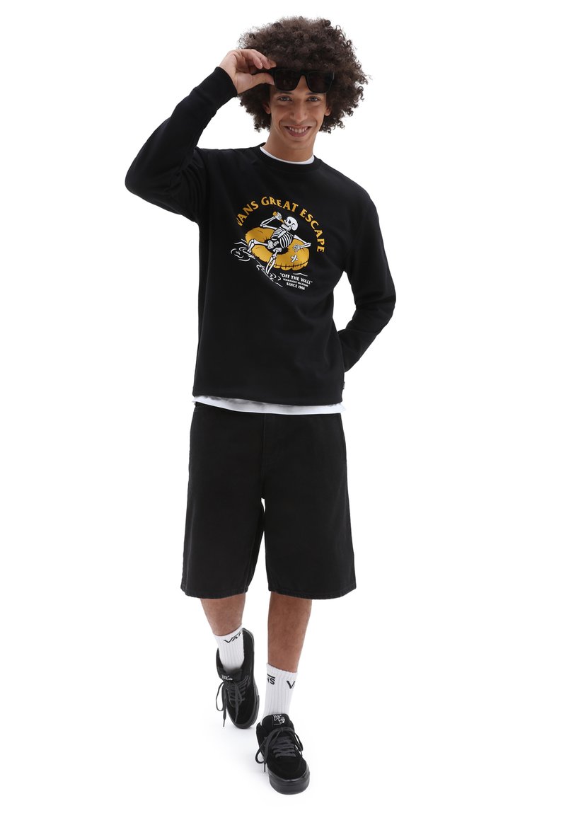 Sudadera negra con un estampado gráfico, combinada con pantalones cortos negros. El modelo lleva zapatillas negras y calcetines blancos. Cabello ligero y texturizado visible.