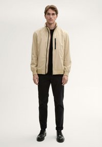 Beige lichtgewicht jas met een staande kraag, voor rits en een ritszak. Draagt over een zwart shirt en gecombineerd met zwarte broek en schoenen.