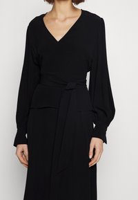 Abito avvolgente nero con scollatura a V, maniche lunghe a sbuffo e cintura annodata in vita. Tessuto liscio con una leggera lucentezza.