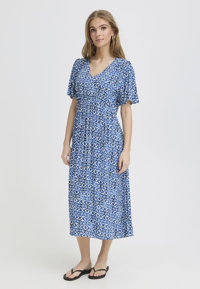 Frau trägt ein blau gemustertes Midikleid mit kurzen Ärmeln und schwarzen Flip-Flops, steht vor einem einfarbigen hellen Hintergrund.