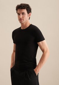 T-shirt noire à manches courtes, col rond, coupe ajustée, texture lisse, porté avec les mains dans les poches, associé à un pantalon noir.