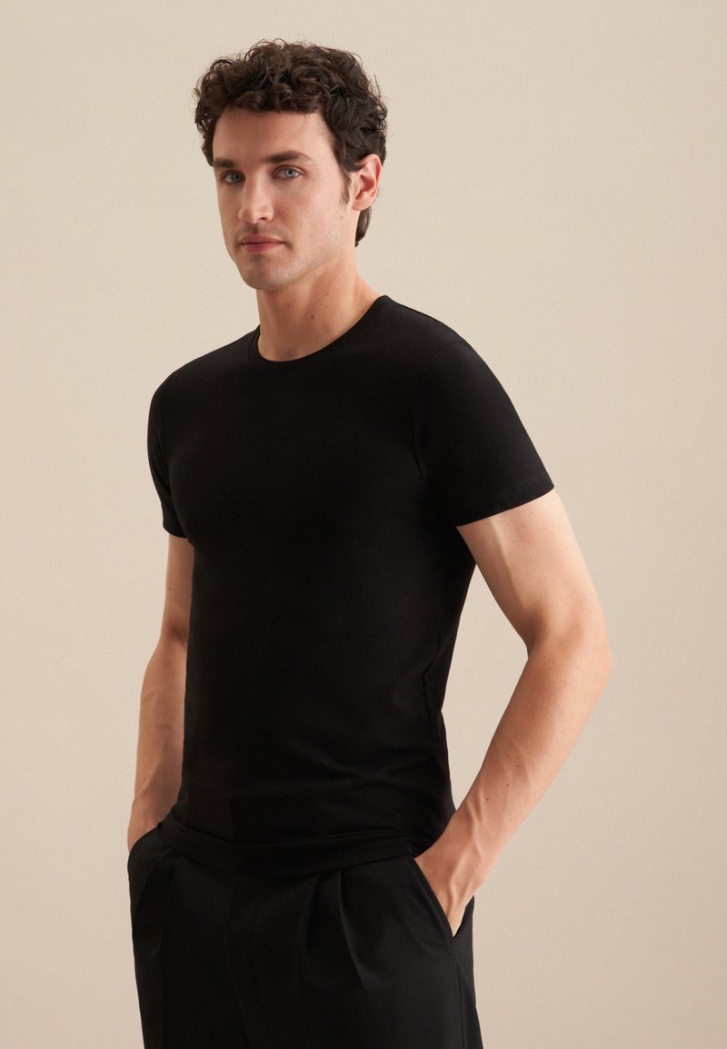 T-shirt noire à manches courtes, col rond, coupe ajustée, texture lisse, porté avec les mains dans les poches, associé à un pantalon noir.