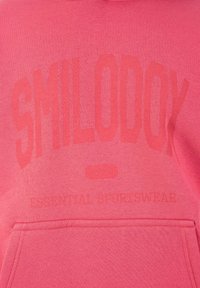 Różowy materiał z wytłoczonym napisem "SMILE-OOCK" oraz mniejszym tekstem "ESSENTIAL SPORTSWEAR" nad przednią kieszenią.