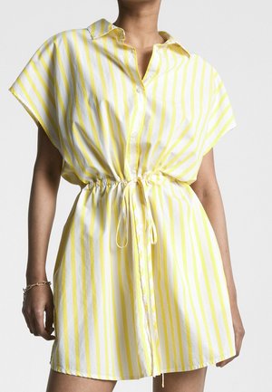 Mujer con un vestido de manga corta a rayas verticales amarillas y blancas, con cuello, botones frontales y cinturilla con cordón.