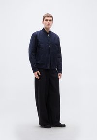 Μπουφάν bomber σε ναυτικό μπλε με μπροστινό φερμουάρ, ριμπ μανσέτες και γιακά. Φοριέται πάνω από φαρδιά μαύρα παντελόνια, δημιουργώντας μια στρωμένη σιλουέτα. Λείο ύφασμα.