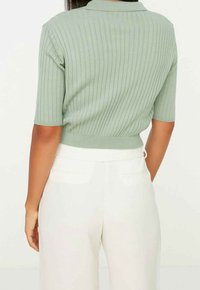 Polo vert en maille côtelée avec manches courtes et ourlet ajusté, associé à un pantalon blanc taille haute. Détail de surface texturée.