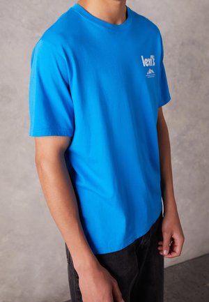Person trägt ein leuchtend blaues Levi's T-Shirt mit kleinem weißem Logo auf der Brust, kombiniert mit dunkler Hose, steht vor einem schlichten Hintergrund.